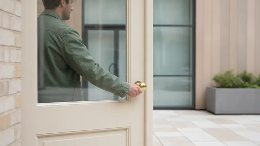 Someone in a green jacket holding open a glass front door on a bright day, seen from outside.